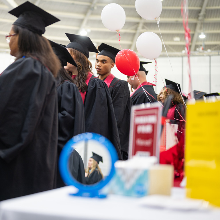Amongst a crowd of graduates, a single graduate in profile is reflected in a hand mirror lying on a nearby table