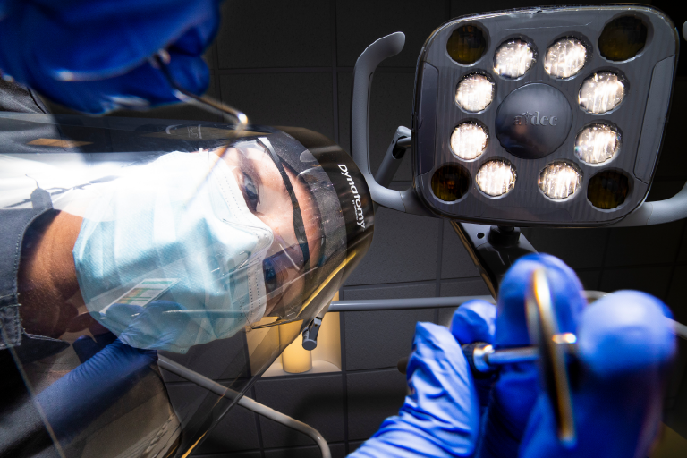 A person wearing a medical mask and shield leans over the camera with a medical device in their hand.
